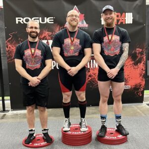 Three powerlifting champions standing on a podium, each holding their medals. They are wearing weightlifting suits and have smiles of accomplishment on their faces.