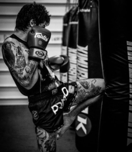 A Muay Thai fighter from Infinite Martial Arts & Fitness throws powerful knee strikes on the heavy bag, dressed in traditional Muay Thai shorts and gloves. The focus and intensity in his eyes show his dedication to his training.