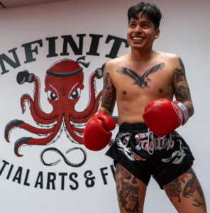 A Muay Thai fighter from Infinite Martial Arts & Fitness, wearing gloves and covered in tattoos, flashes a victorious smile after a grueling training session. The sense of accomplishment and camaraderie is evident in his expression