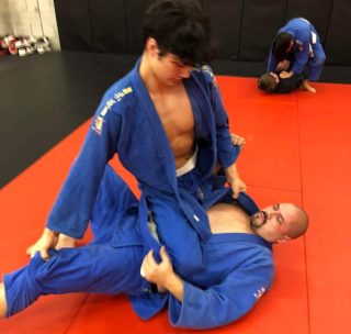 Young man in blue gi training Brazilian Jiu-Jitsu in mounted position on a larger man in blue gi