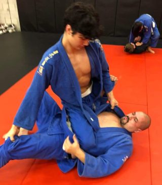 Young man in blue gi training Brazilian Jiu-Jitsu in mounted position on a larger man in blue gi